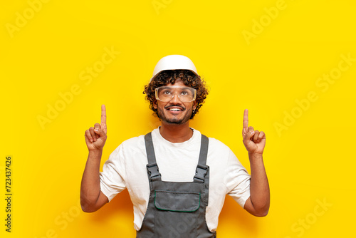 young indian male builder in hard hat and overalls points with his hands up on a yellow isolated background, indian foreman in uniform raises his hands and shows and advertises mine space photo