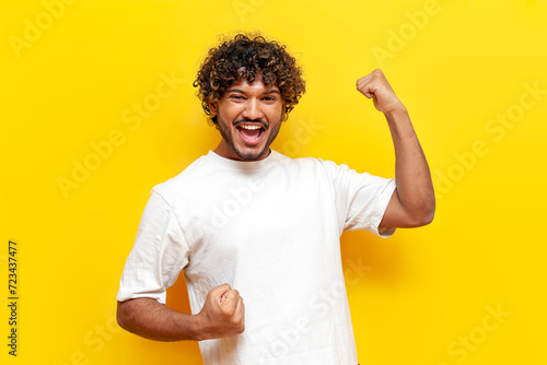 cheerful indian man celebrating victory and good luck with raised hands on yellow isolated background, curly guy wins and rejoices at success photo
