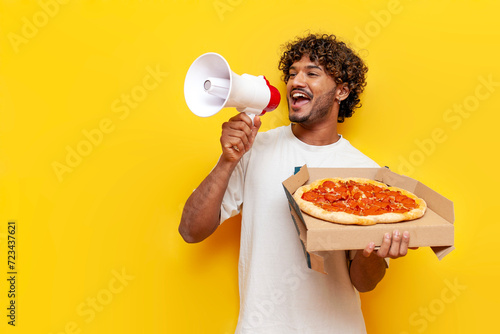 young Indian man holding a delicious pizza and announcing information into a loudspeaker on a yellow isolated background, a curly-haired guy shouts into a megaphone and advertises fast food