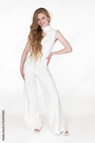 Happiness young woman stylishly dressed in white long jumpsuit straight design standing in full length. Caucasian woman with long blonde hair looking at camera. Studio shot on white background © Alexander Piragis