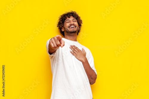 young indian man in a white t-shirt mocks and jokes and points his finger forward on a yellow isolated background, curly-haired guy laughs and ridicules and mocks photo