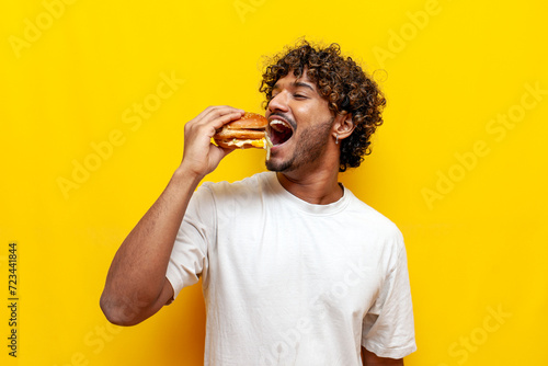 young indian man eating a big tasty burger with his mouth wide open on a yellow isolated background  curly guy student eating and advertising fast food