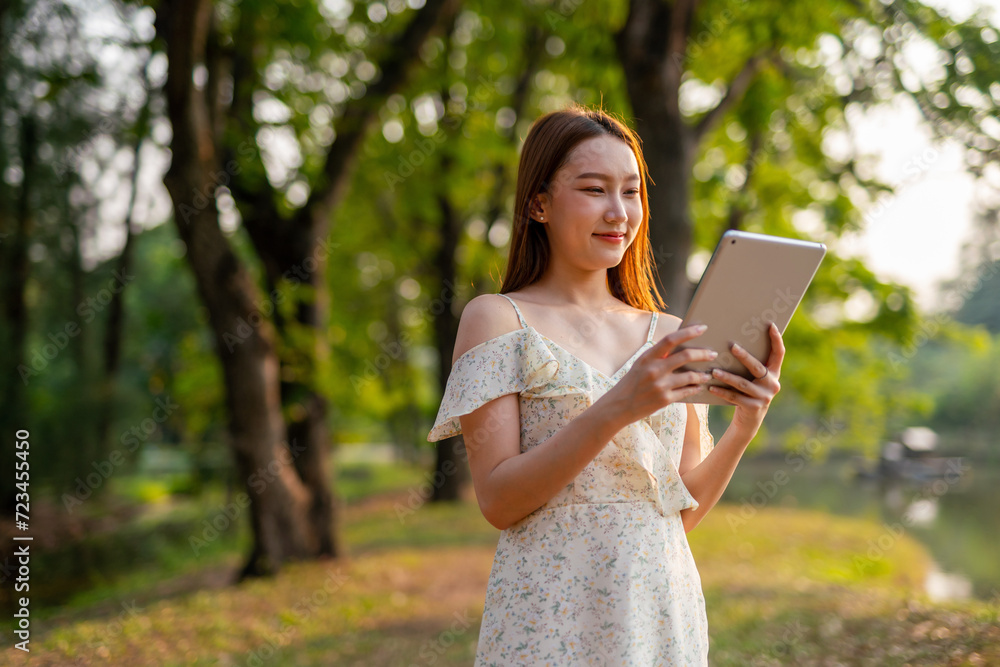Naklejka premium Beautiful Young Asian Woman Finds Serenity, Nature, and Inspiration While Holding a Digital Tablet in the Sunset Breeze at the Park