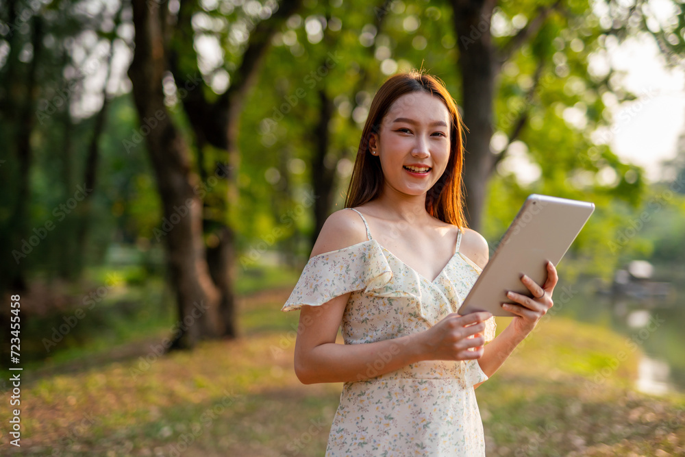 Naklejka premium Beautiful Young Asian Woman Finds Serenity, Nature, and Inspiration While Holding a Digital Tablet in the Sunset Breeze at the Park