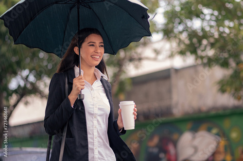  An executive woman with a black jacket walks down the street on a rainy day holding an umbrella and a coffee in her hand.