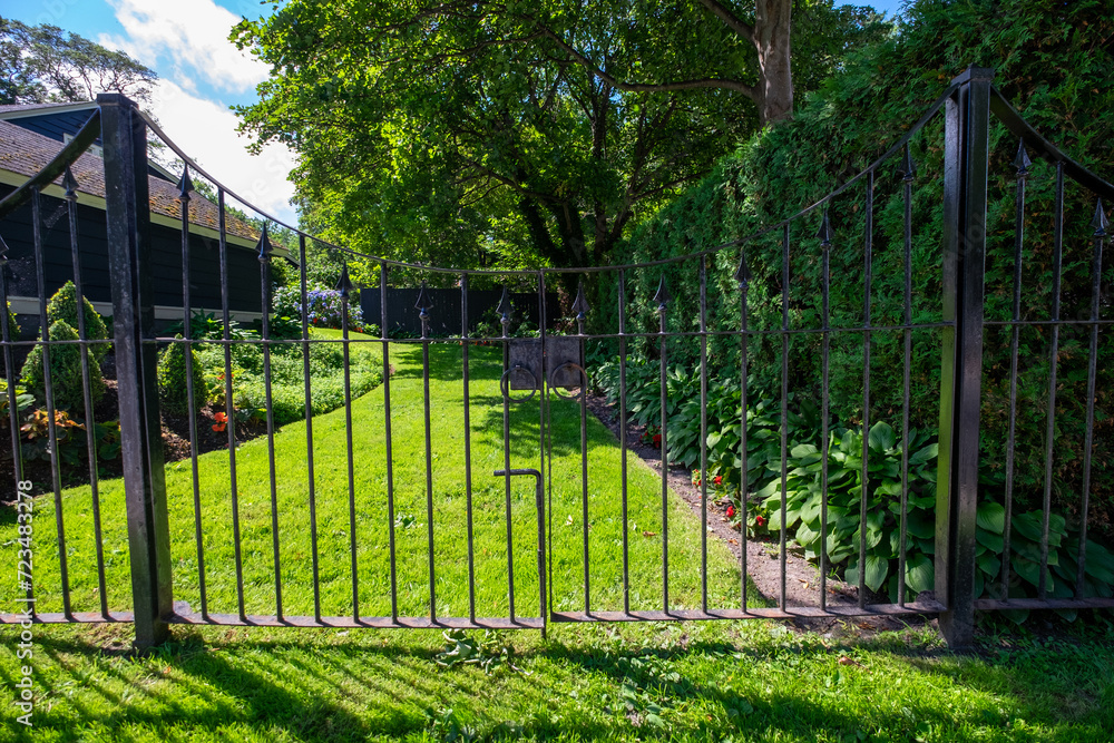 A closed black wrought iron fence and gate with lush green grass, rows of flowers, shrubs, and maple trees. A blue wooden building with white trim in the yard under a blue sky with white clouds. A closed black wrought iron fence and gate with lush green grass, rows of flowers, shrubs, and maple trees. A blue wooden building with white trim in the yard under a blue sky with white clouds.
