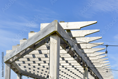 A grey colored worn and weathered wood pergola roof with a blue sky in the background. The outdoor sun shelter has wooden slats hanging over the wooden beams with string lights hanging off the boards.