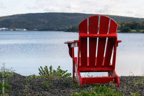 A bright empty red Adirondack chair on the edge of a green grassy meadow overlooking the blue ocean with mountains covered in trees, a bay, and a cove. The water is flat calm and the sky is cloudy. 