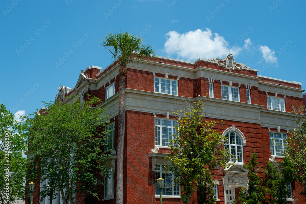 A multi-level vintage red brick building with cement ledges along the top two floors. There are glass windows with white trim. There are tall lush green trees around the exterior wall. The sky is blue
