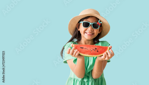 Happy Asian little girl posing with wear a hat with sunglasses holding watermelon slices, Holiday summer fashion green dress, isolated on pastel blue color background photo