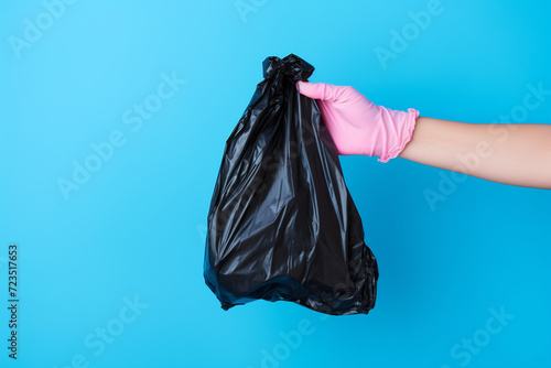 Hand in Pink Glove Holding a Black Garbage Bag Against a Blue Background. Waste Management Concept