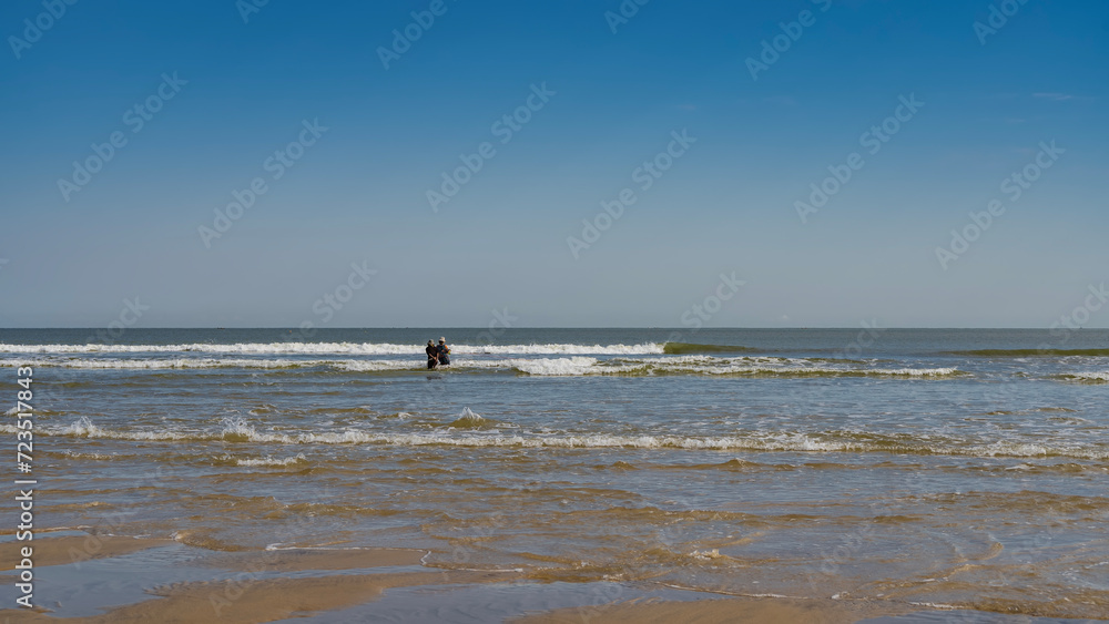 The waves of the turquoise ocean roll towards the shore, foaming. Puddles of water on a sandy beach. Two people are standing knee-deep in the sea, pulling out a fishing net. Clear blue sky. Copy space
