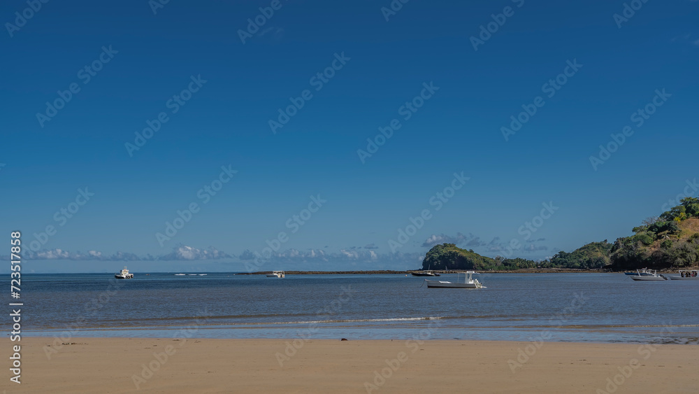 The boats are anchored in the calm blue ocean. Waves roll onto a sandy beach. A green hill against a clear azure sky. Light clouds over the horizon. Copy space. Madagascar. Nosy Be 