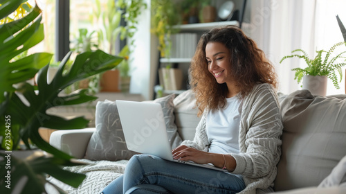 Office at home girl at laptop. Young woman, using laptop remote working at home office looking at computer communicating by video call.He is sitting on the sofa against a background of plants.