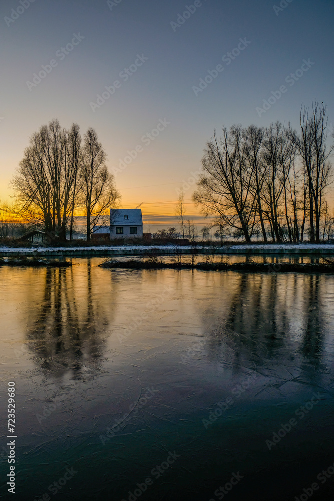This tranquil image encapsulates the serene beauty of a riverside cottage at dawn. The first light of the day casts a soft, golden glow on the scene, with the frozen river in the foreground reflecting This tranquil image encapsulates the serene beauty of a riverside cottage at dawn. The first light of the day casts a soft, golden glow on the scene, with the frozen river in the foreground reflecting