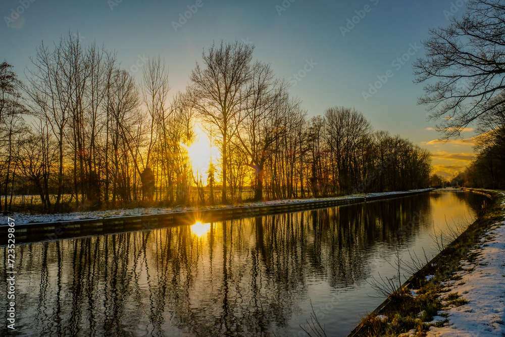 This image depicts a stunning sunset scene where the sun's golden rays pierce through the silhouetted trees along a calm canal. The sky, ablaze with warm tones, transitions from yellow to a deep blue