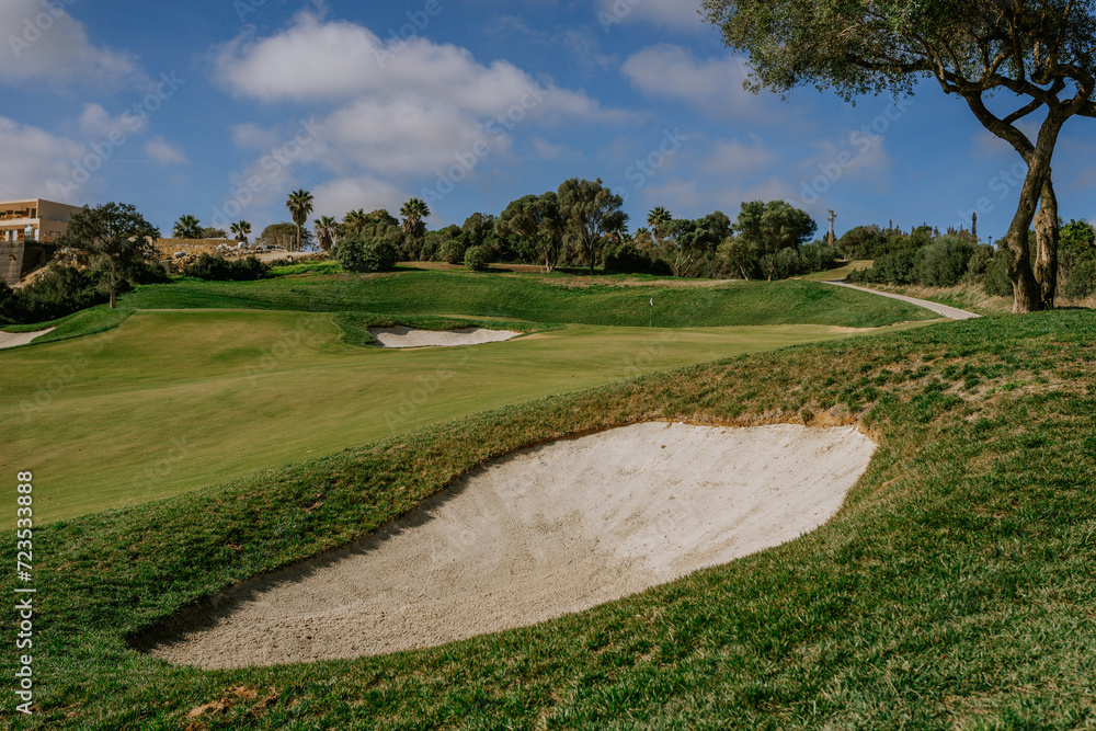Sotogrande, Spain - January, 23, 2024 - Golf course with sand bunker, green grass, trees, and pathway under a partly cloudy sky.