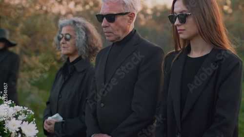 Tilt side footage of close relatives in black clothes and sunglasses standing near wooden casket with white flowers on lid during funeral service outdoors
