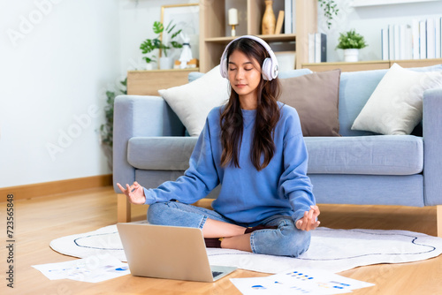 Happy young Asian woman practicing yoga and meditation at home sitting on floor in living room in lotus position and relaxing with closed eyes. Mindful meditation and wellbeing concept photo