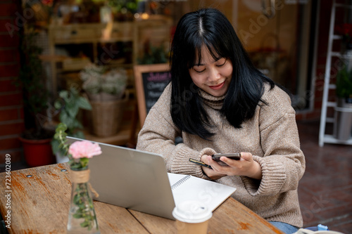 Positive Asian woman is responding to messages on her phone while working remotely at a coffee shop.