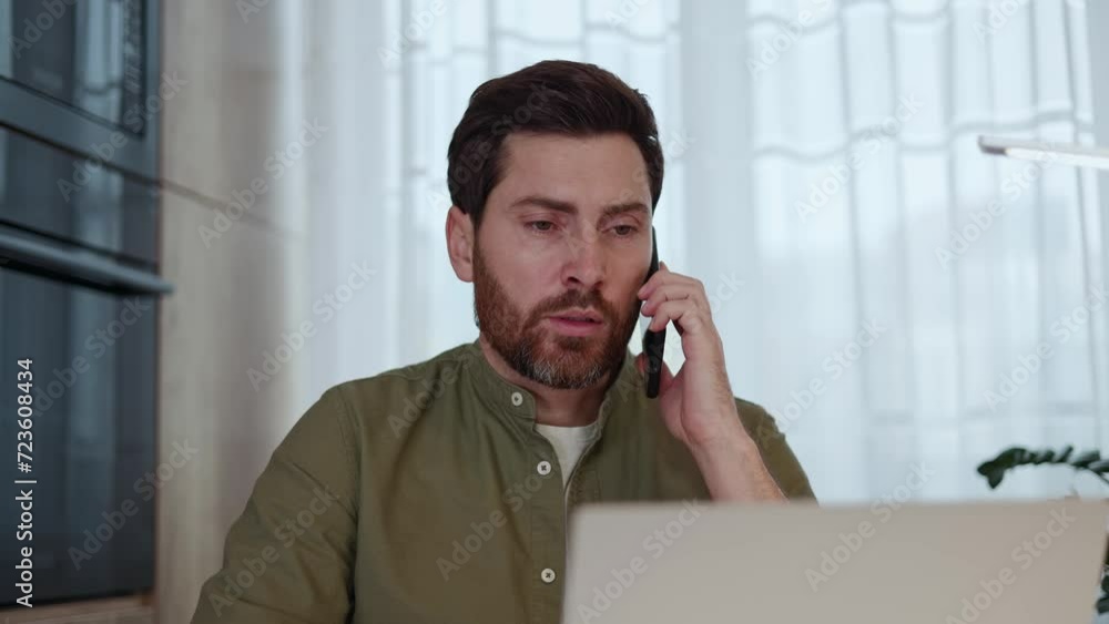 Concerned man with fancy hairstyle picking up smartphone call while sitting by desk with portable computer. Handsome remote worker having serious discussion and smiling while looking at monitor. Concerned man with fancy hairstyle picking up smartphone call while sitting by desk with portable computer. Handsome remote worker having serious discussion and smiling while looking at monitor.