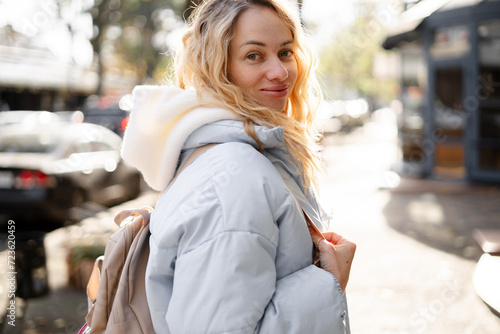 Young stylish woman wearing blue jacket, white pants with backpack walking in the city street in cold season. Winter fashion, outfit in pastel colors. Small size model.