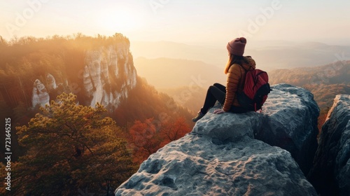 Girl sitting on the top of mounting and enjoying yellow sunrise above sea. Hiking woman in khaki jacket relaxing on the cliff looking at a beautiful sunlit landscape. Green valley in sunlight