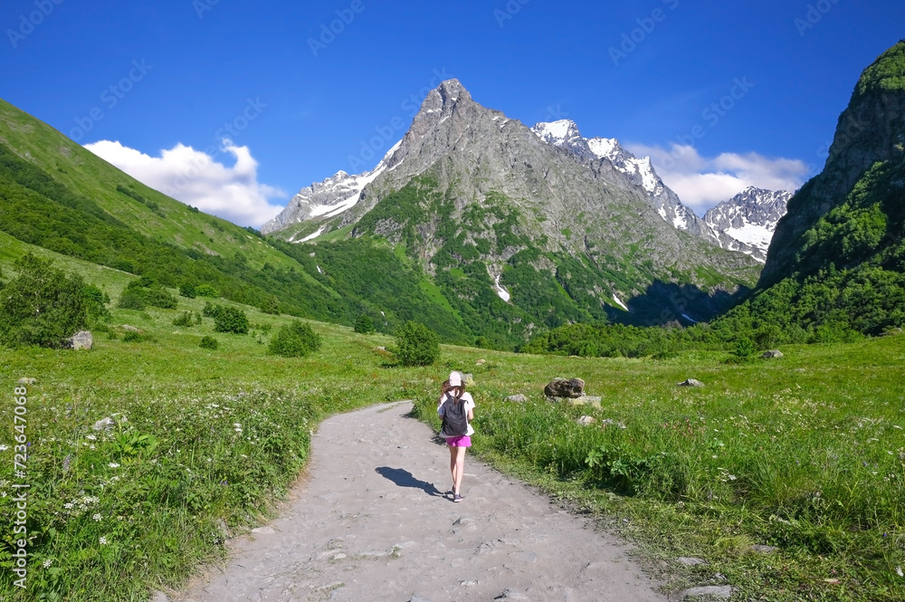 Tourist girl with a backpack walks along a dirt road among mountains with peaks covered with snow