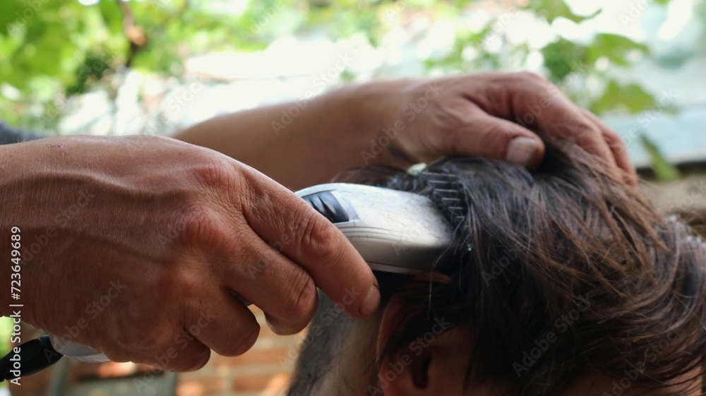 Fototapeta premium hands of a hairdresser cutting the back of a person's head with a clipper, close-up, shaving long dark hair from the back and top of the head with a trimmer with an attachment