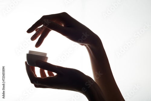 Close-up of a mockup of a white jar with moisturizer in a woman hand on a white background. A woman uses cream from a jar, smearing it over her hand.