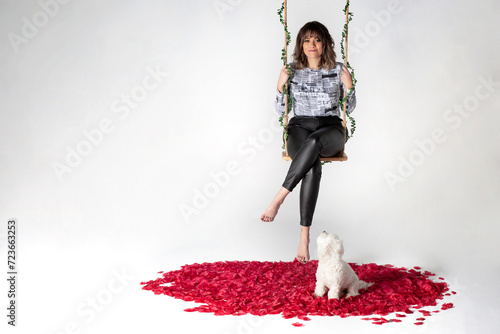 Chica morena con mechas y pelo rizado columpiándose en un columpio de madera con hojas sobre fondo blanco el día de san valentín con su perro adorable bichón blanco. Suelo con pétalos de rosa