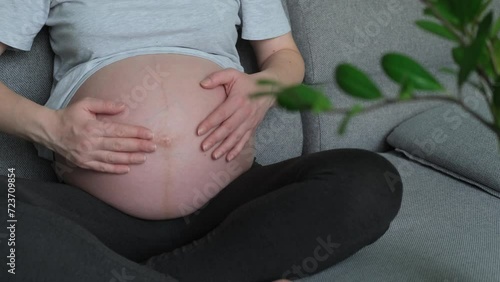 4k.A pregnant girl gently strokes her belly while sitting on the sofa. Waiting for the birth of a child. Woman hands touching her tummy, waiting child. Preparation for childbirth. Happy motherhood.