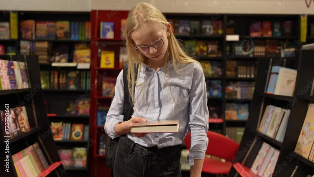 Young school girl standing in the bookstore between shelves and reads books description. Smart, educated blond hair girl. Young school girl standing in the bookstore between shelves and reads books description. Smart, educated blond hair girl.