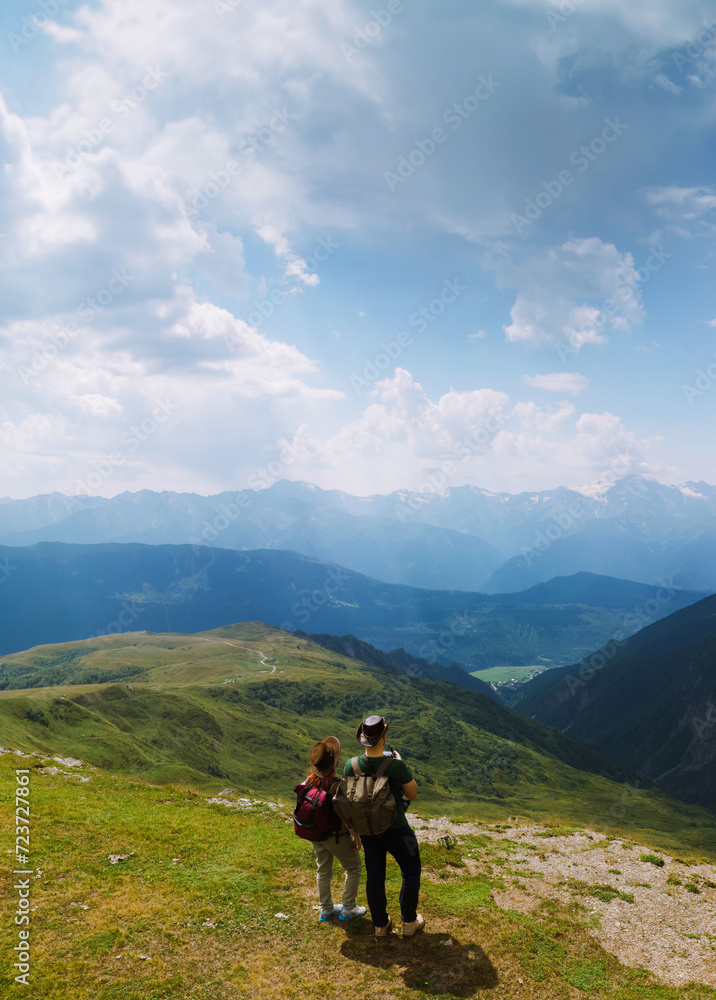 Couple woman and man wearing wide sun hat and backpack, tourists on trail to Koruldi Lakes, Mestia Georgia. The concept of travel and active recreation. Summer day. Aerial vertical photo