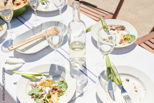 The remains of a summer feast lay on a sunlit table, with empty dishes, scattered crumbs, and half-empty glasses of white wine, capturing the relaxed end of a joyful meal.