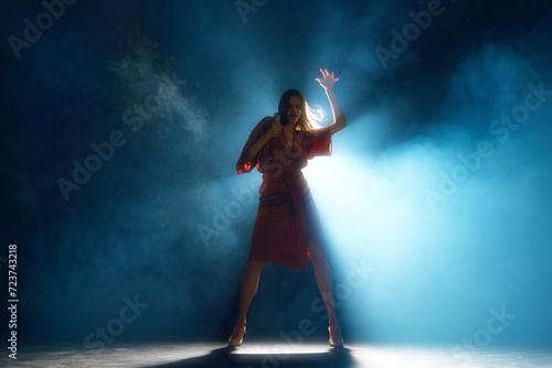 Woman, vocalist in moment of passion, highlighted by backlight and theatrical smoke on stage against black background. Concept of hobby, festival, concert, disco, entertainment. Ad