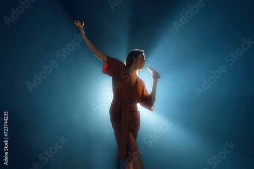 Graceful singer with uplifted arm, bathed in serene blue spotlight on stage against dark background and theatrical smoke. Concept of hobby, festival, concert, disco, music and dance. Ad