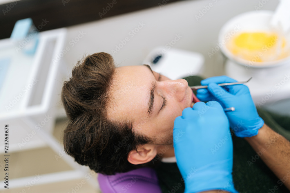 Closeup face of male patient sitting in stomatology seat with open mouth during unrecognizable male dentist examining teeth, with dental tool in dentistry clinic. Concept of stomatology. Closeup face of male patient sitting in stomatology seat with open mouth during unrecognizable male dentist examining teeth, with dental tool in dentistry clinic. Concept of stomatology.