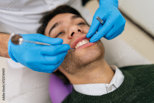 Closeup face of young man patient at dentist chair during dental procedure. Close-up hands of unrecognizable doctor examining patient teeth in modern clinic. Healthy teeth and medicine concept.