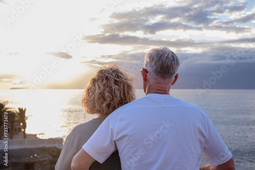 Rear view of mature and romantic senior couple face the sea looking at horizon over water, two smiling people stay together expressing love and tenderness