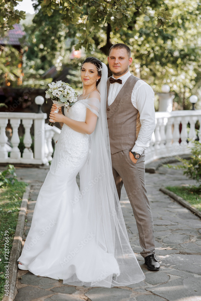 A handsome groom embraces his bride in a lush white dress and smiles in a beautiful outdoor setting. Under the open sky. High quality photo. A newlywed couple poses together on a sunny summer day.