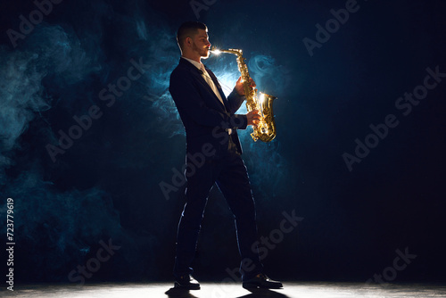Solo saxophonist in concert outfit performing with intense expression on stage with backlight against dark background with dramatic smoke. Concept of art, instrumental music, dance, culture. Ad