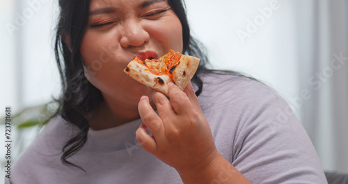A chubby Asian woman enjoying pizza while relaxing on the sofa at home.