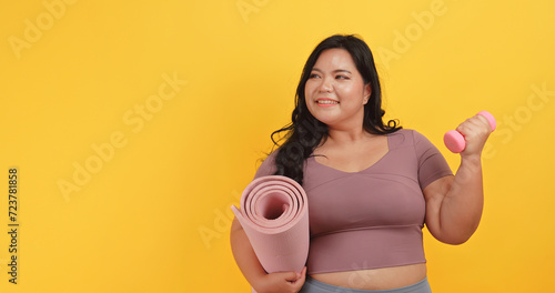 A chubby Asian woman in sportswear is holding dumbbell and yoga mat on yellow background.