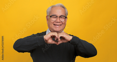 An old Asian man making a heart shape with his hands. Isolated on yellow background in the studio.