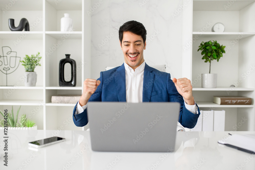 A jovial Hispanic businessman celebrates success or good news on his laptop in a sleek, contemporary office, exuding confidence and achievement. Concept of positive results in business