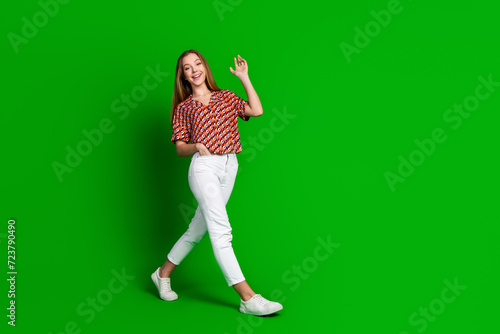 Full size photo of nice lovely teen girl wear print blouse white trousers say hi going to empty space isolated on green color background © deagreez