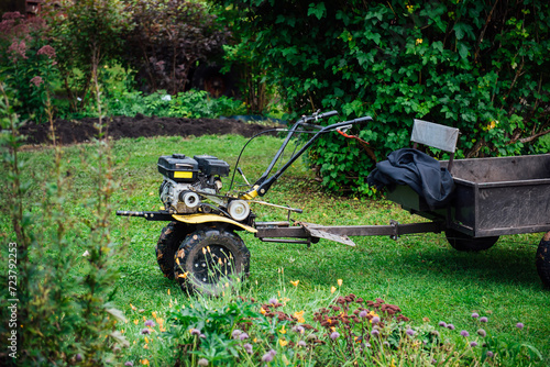A mini hand-held tillerblock with a cart trolley for working in the garden and on the farm against the background of green bushes and lawns. Rural life, gardening and farm work