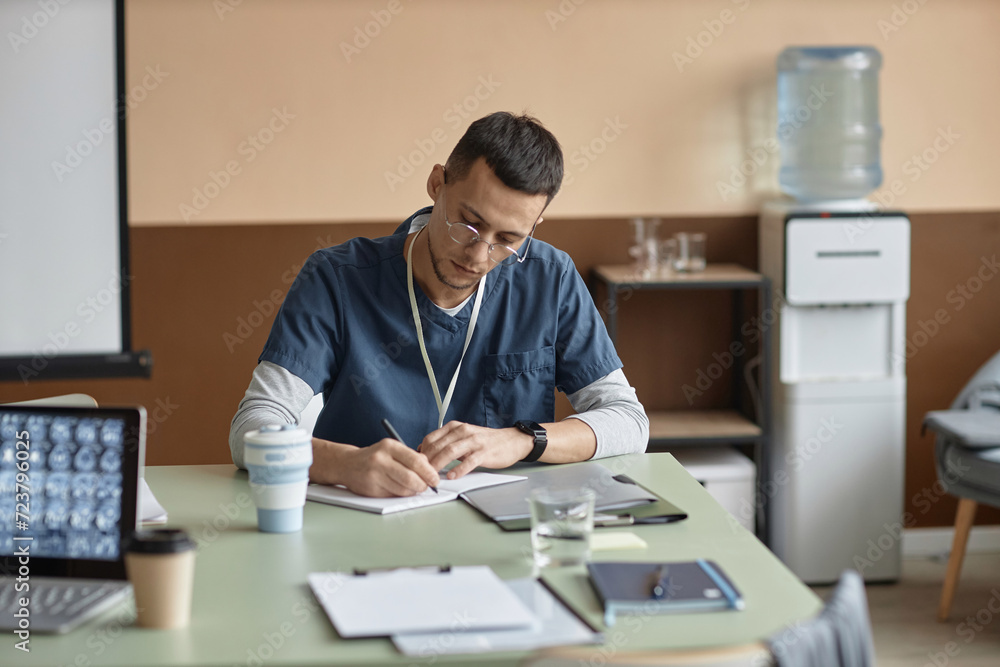 Medium shot of young Islamic man doctor wearing eyeglasses writing in notebook sitting at table in conference room Medium shot of young Islamic man doctor wearing eyeglasses writing in notebook sitting at table in conference room