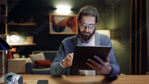 Carefree millennial man in eyewear relaxing at home in evening with digital touchpad in hands, searching information, typing message, communicating distantly in social network, shopping. photo
